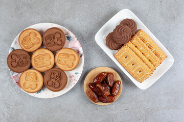 Two plates of cookies and biscuits next to a small wooden tray of dates on marble background
