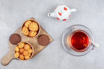 Cookies in bowls and on wooden board, with a cup of tea and a small teapot on marble background