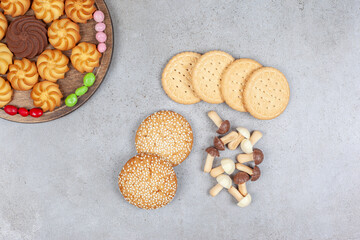Small piles of cookies and chocolate mushrooms next toa plate of various cookies encircled by candies on wooden board, on marble background