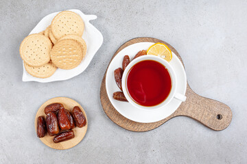 A platter of cookies and a small pile of dates next to a cup of tea with some dates on saucer on a wooden board, on marble background