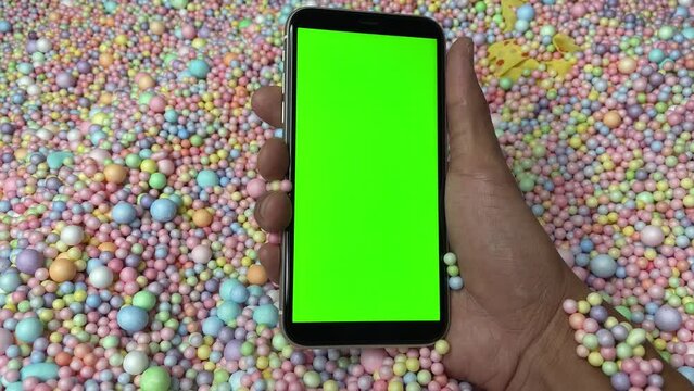 Chroma key green screen on smartphone vertical in male hand with colourful pastel rainbow foam ball beads in background