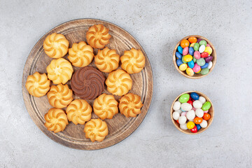 Two bowls of candy next to a decorative arrangement of cookies on wooden board on marble background