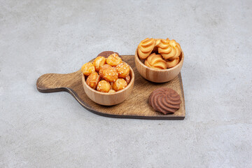 Two bowls of cookies next to brown cookies on wooden board on marble background