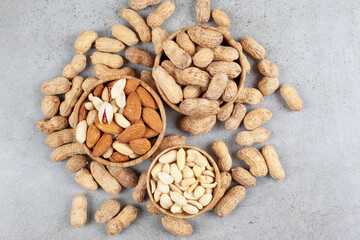 A pile of different nut types in bowls next to scattered peanuts on marble background