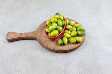 Bowl of kumquats next to a pile on a wooden board in marble background