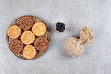 Decorated cookies on wooden board next to a sack and a small bowl of mullberries on marble background