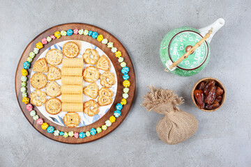 A teapot, a sack and a small bowl of dates next to a plate of cookies encircled by popcorn candy on wooden board on marble background
