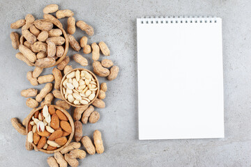 Bowls of assorted nuts and scattered peanuts next to a notebook on marble background