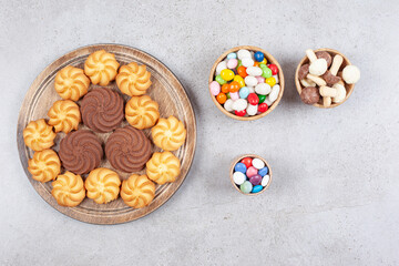 Cookies on wooden board next to bowls of candies and mushroom chocolate on marble background