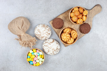 Bowls of cookies next to brown cookies on wooden board with russian sweets, a sack and a bowl of candy on marble background