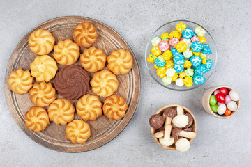 Cookies on wooden board next to bowls of candies and mushroom chocolate on marble background