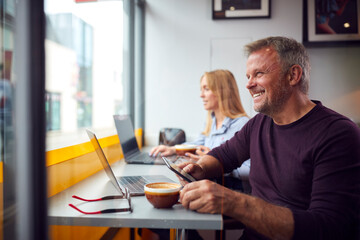 Customers In Coffee Shop Window Working On Laptops Using Mobile Phones