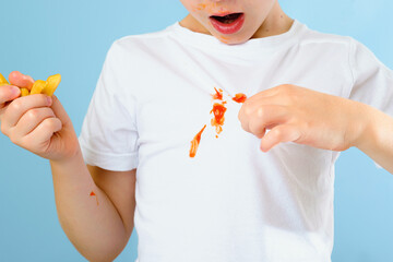 Surprised boy showing dirty stain from ketchup sauce on a white shirt. Isolated on blue background. A child holding French fries in his hand. daily life dirty stain concept