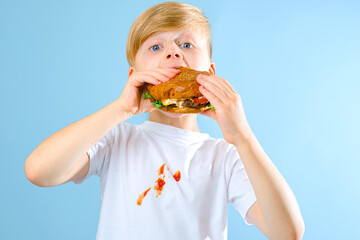 Portrait of a boy biting hamburger and looking at the camera. isolated on blue background. Dirty stain tomato sauce on white t-shirt. daily life dirty stain concept © HalynaRom