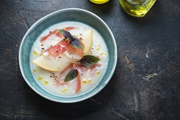 Bowl of cold melon soup served with jamon, above view on a dark-brown stone background, horizontal shot with space