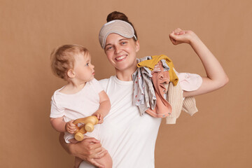 Horizontal shot of smiling woman with dark hair wearing white t shirt and blindfold standing with her baby daughter, stretching her arm, waking up in good mood, posing isolated over brown background.