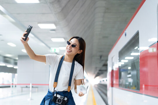 Selective focus half-body image of a happy Asian female traveler with a film camera, wearing sunglasses, standing on a subway platform using a cellphone taking selfies with a blurred stationary train.