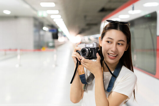 Selective focus and close-up of a pretty Asian woman with sunglasses on head, smiling holding a vintage film camera with two hand prepare to take a picture in front of her at a blurred subway station.