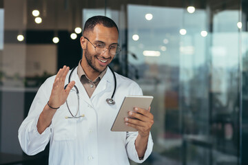 Cheerful hispanic doctor in medical white coat and stethoscope using tablet computer for video call and online consultation to patients, man working inside clinic office building.