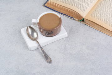 Cup of milk coffee, spoon and book on stone surface