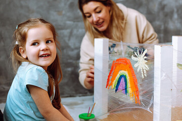 Little smiling blond girl painting rainbow on adhesive tape with teacher. Having fun and learning...