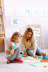 Vertical passionate female teacher play and paint with little girl on cardboard, learn numbers in kid development school