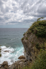 Vertical photo shows a rocky with vegetation. Wavy sea and sky with clouds at he background.