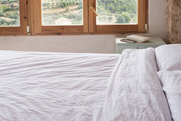 Interior habitación con ventana con vistas a la naturaleza, cama con sábanas blancas, nordico. En la mesita libros y movil