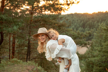 Adorable, cheerful, laughing woman, mother in hat carrying on back tired little girl, daughter on...