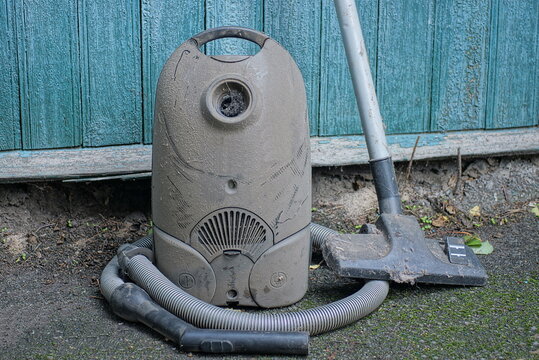 one old dirty black vacuum cleaner in gray dust with hose and brush on tube stands on the asphalt near the green wall on the street