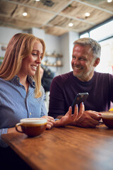Couple Sitting At Table In Coffee Shop Looking At Mobile Phone Together