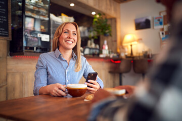 Couple Sitting At Table In Coffee Shop Looking At Mobile Phone Together