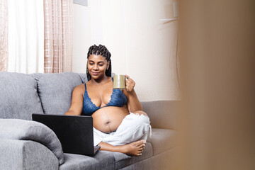 Young black pregnant woman sitting holding a cup using a laptop.
