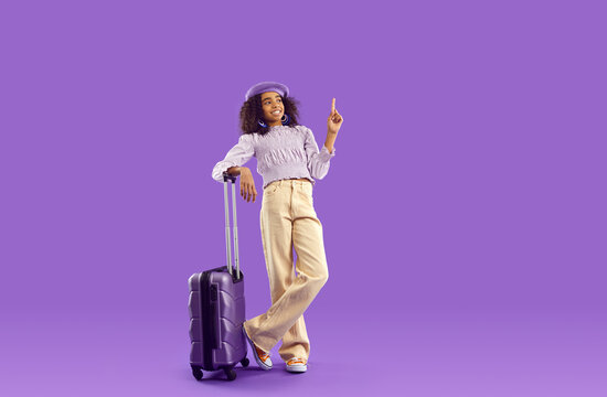 Joyful African American girl teenager with travel suitcase showing up recommending to pay attention to copy space dressed in casual clothes stands in purple studio. Tourism, holidays concept