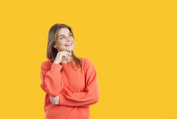 Happy young woman thinking with hand on chin and positive expression. Creative cheerful confident modern teenage girl in orange jumper dreaming of something good on vivid yellow copyspace background