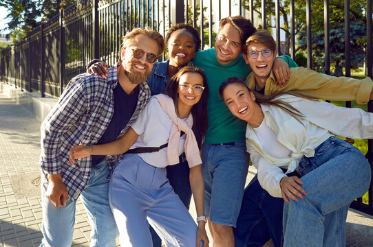 Portrait of cheerful group of youth best friends having fun posing for photo while walking on city streets. Multiracial men and women in stylish casual streetwear are hugging and smiling at camera.