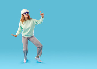 Fototapeta na wymiar Full body length shot of funny happy pretty young woman in pastel mint sweatshirt, grey trousers, white faux fur hat and cool sunglasses dancing isolated on blue copy space background. Fashion concept