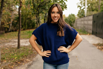 Happy exited lovely girl with wonderful smile wearing blue knitted shirt posing at camera in city on background of autumn park un sunny day. 