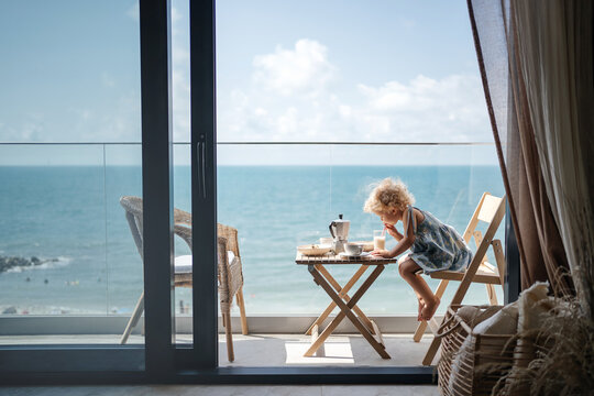 small child girl with blond curly hair has breakfast in the attic overlooking the seascape. Drinks milk.