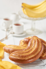 A crescent-shaped plate of fresh buns sits on a white wooden table. In the background are several white cups of tea and a fruit bowl with bananas.