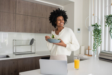 Portrait of young woman working from home having lunch. Happy female model eating salad, standing...
