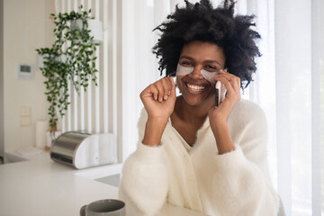 Portrait of young woman with cosmetic eye patches working from home. Happy female model looking at camera, sitting at table, speaking on phone. Portrait, remote work concept