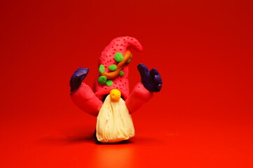 A figure of a dwarf made of plasticine with raised hands on a red background. Christmas figurines.