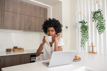 Businesswoman drinking coffee at home and looking at camera. Cheerful young woman sitting at table in kitchen, working on laptop from home, having breakfast. Home office, business, technology concept