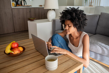Focused businesswoman sitting on floor and typing on laptop. African American copywriter looking at keyboard, chatting with colleague online during self-isolation. Home office, pandemic concept - obrazy, fototapety, plakaty