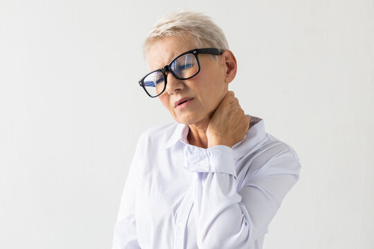 Portrait Of Senior Woman With Severe Pain In Neck. Female Model In White Shirt Touching Neck, Rubbing, Trying Soothing Pain. Portrait, Studio Shot, Pain Concept