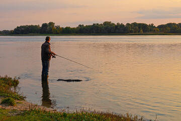 A fisherman stands in the water on the river bank with a fishing rod in his hands in the evening. Fishing with spinning