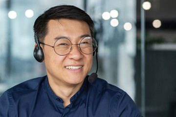 Portrait of man with headset for video call, asian worker smiling and looking at camera, tech customer support and service, businessman working inside modern office building.