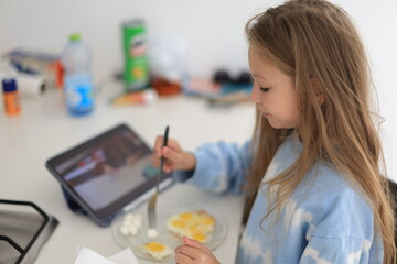 Positive caucasian little girl having breakfast and looking at tablet at home