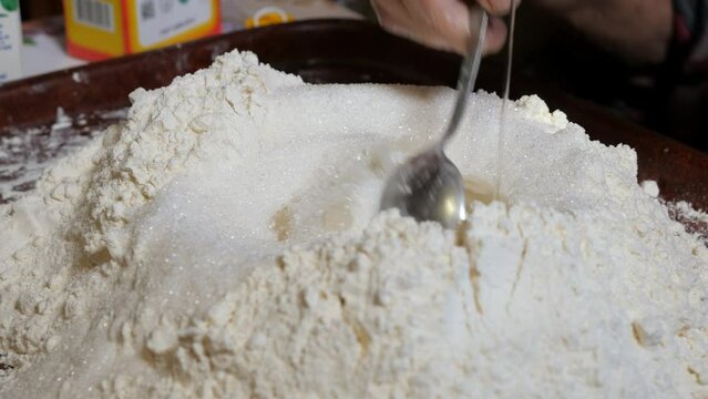 Adding granulated sugar and vegetable oil to flour for baking homemade balls of kolobok. Mixing ingredients with a metal tablespoon.